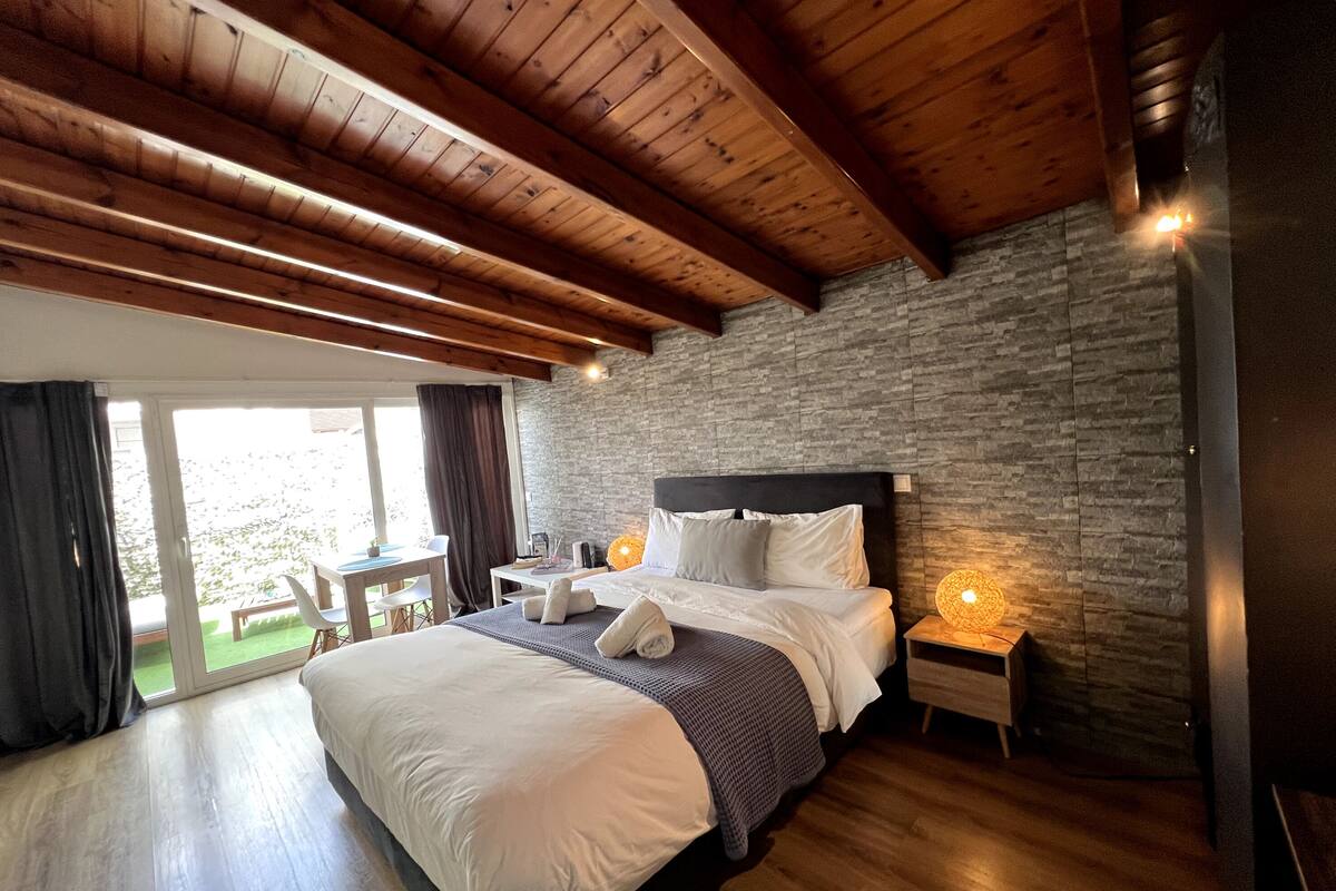 A bedroom with a large bed, wooden ceiling beams, a stone wall, and a small table with chairs.