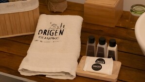 Un baño ordenado con una toalla blanca, un dispensador de pañuelos de madera y tres botellas de champú ORIGEN sobre una bandeja de madera.