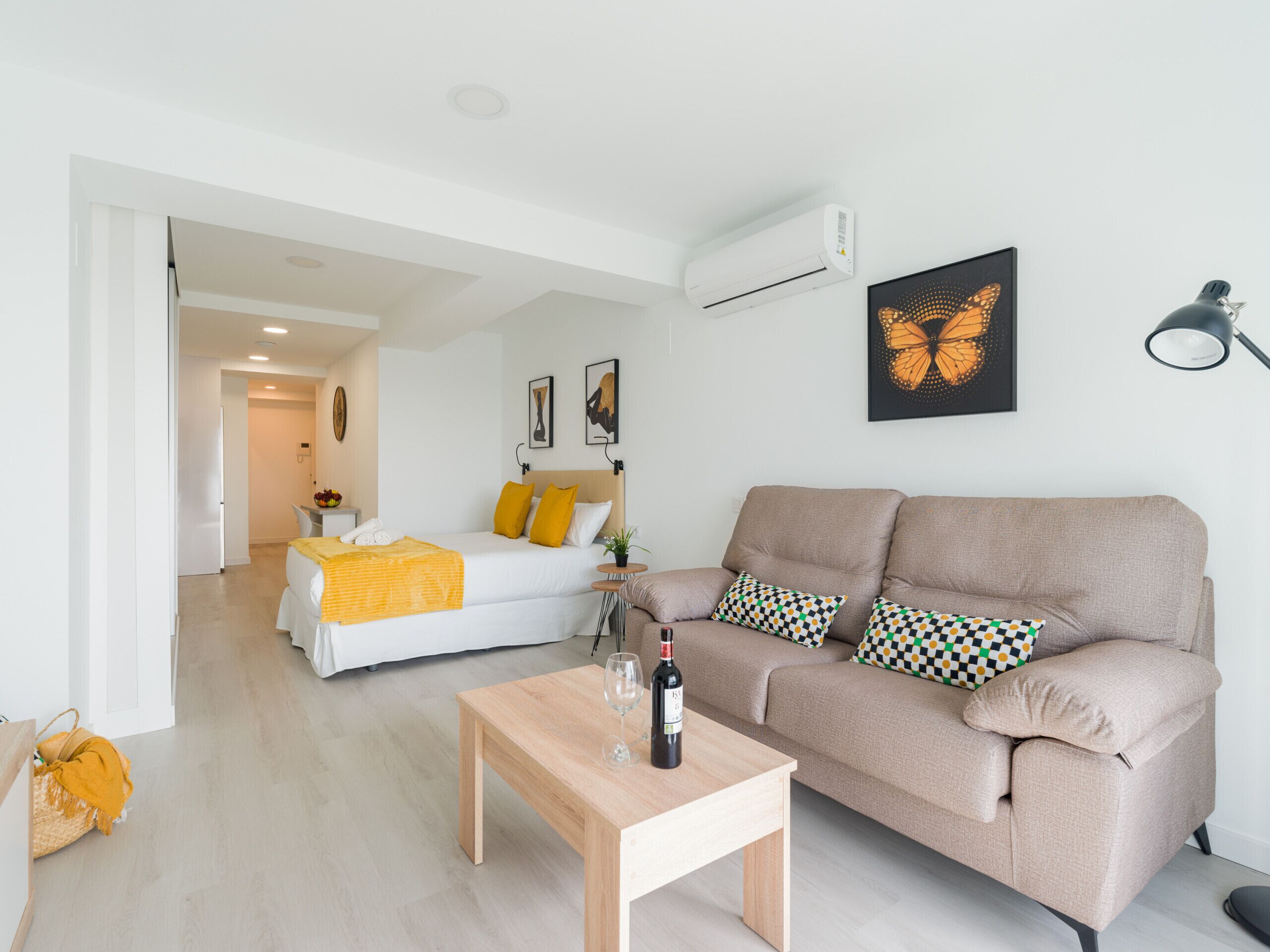 Ein modernes Wohnzimmer mit einer Couch, einem Holztisch und einem Bett mit gelben Kissen. An der Wand hängt ein Bild von einem Schmetterling, und rechts steht eine Lampe.