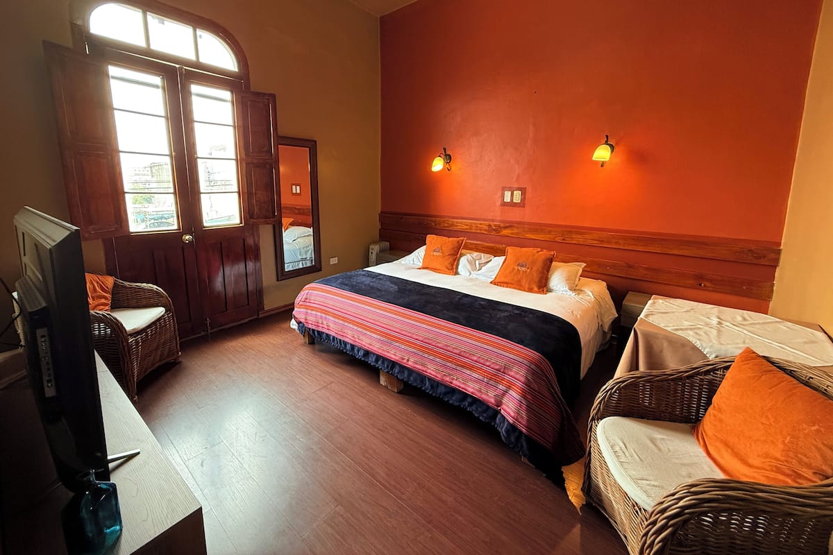 Une chambre d’hôtel avec un grand lit, deux lampes fixées au mur, une tête de lit en bois, un fauteuil en osier, un bureau avec un ordinateur et une fenêtre donnant sur les bâtiments environnants.