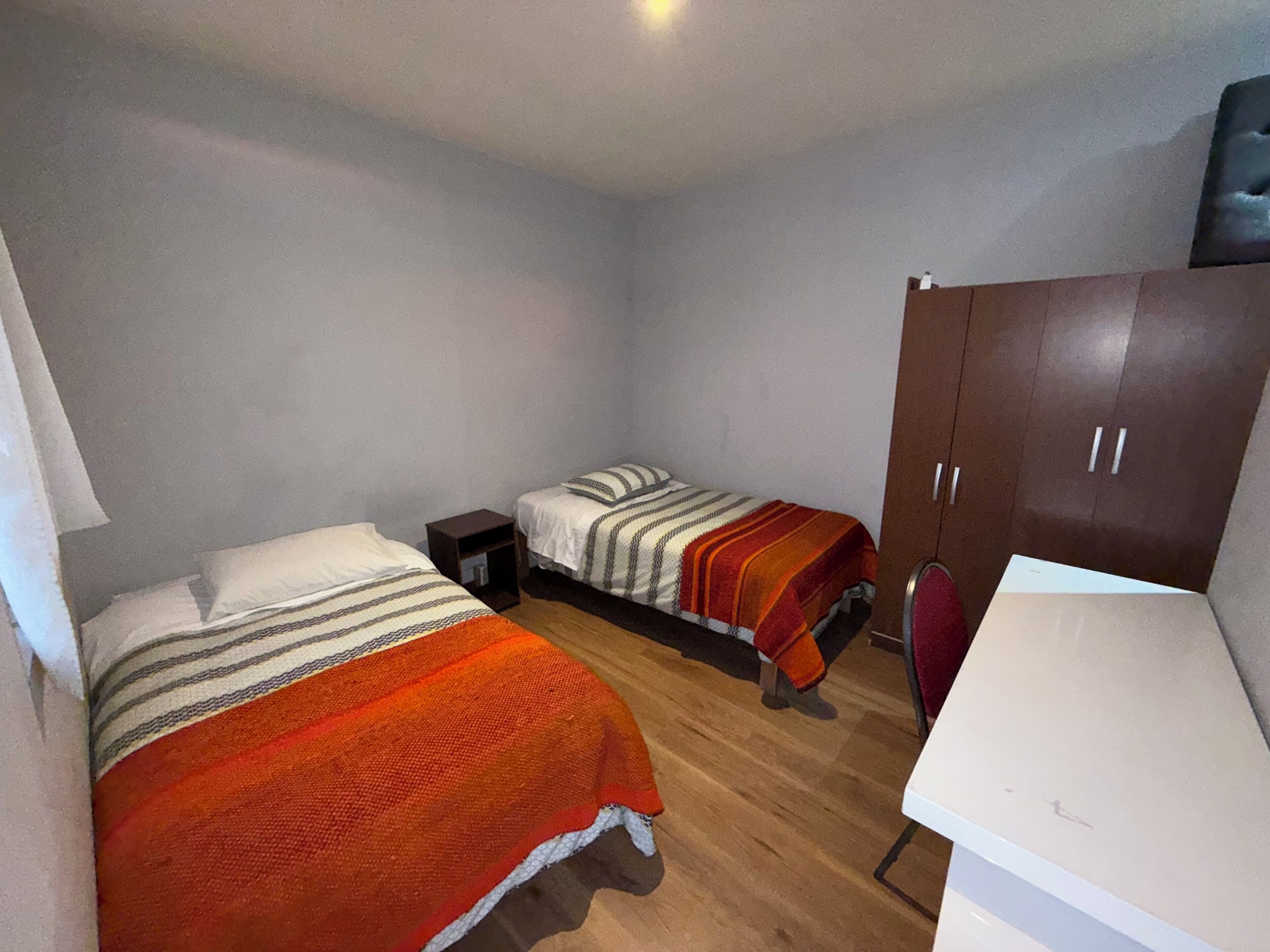 A small, sparsely furnished room with two single beds, a wooden floor, a white desk, and a wardrobe.