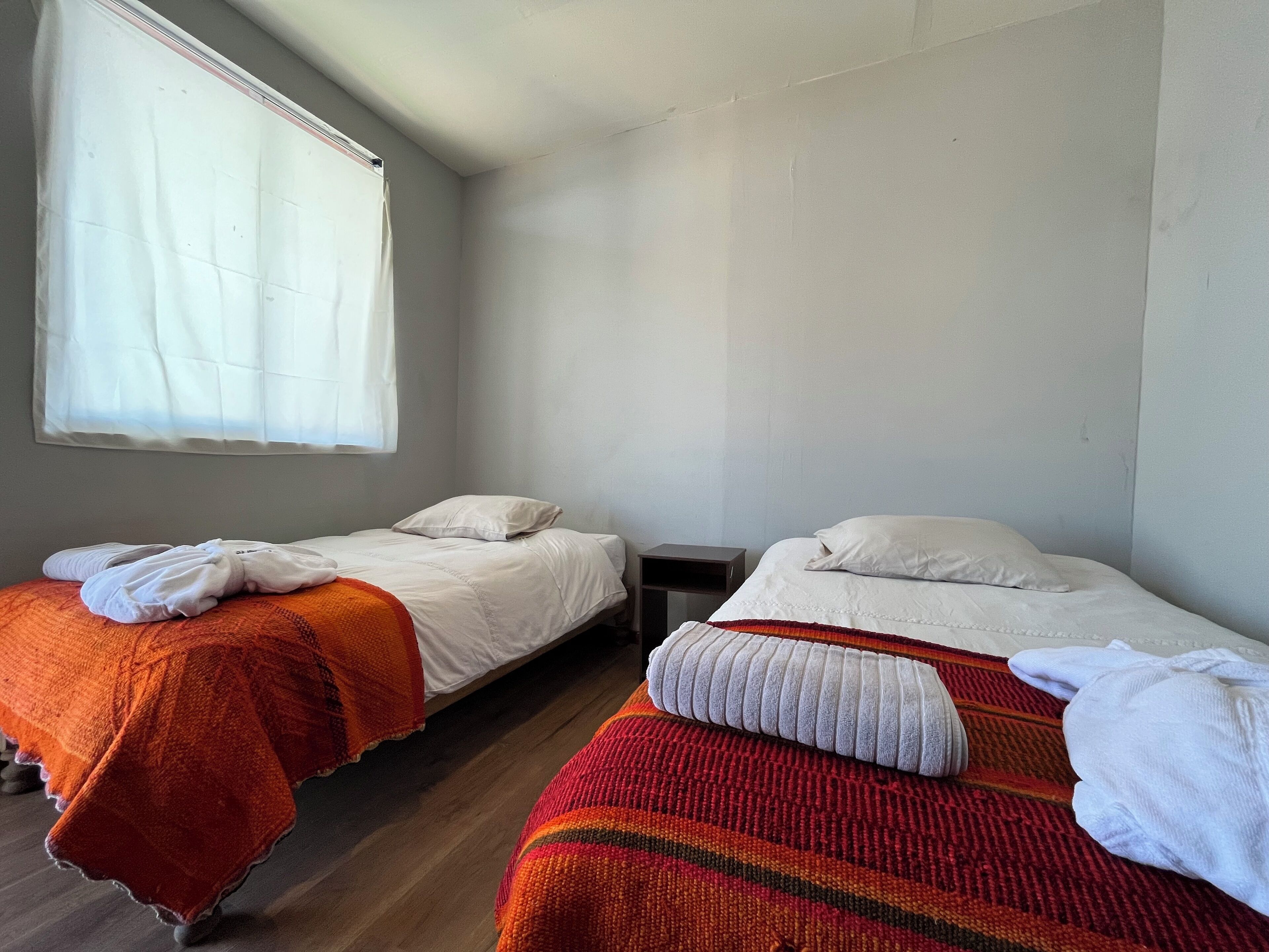 A room with two beds, each with white linens and a colorful blanket. There is a small nightstand between the beds, and a window with white curtains on the left side.