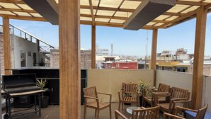 A rooftop patio with wooden chairs and a table, a grill, and a view of the cityscape.