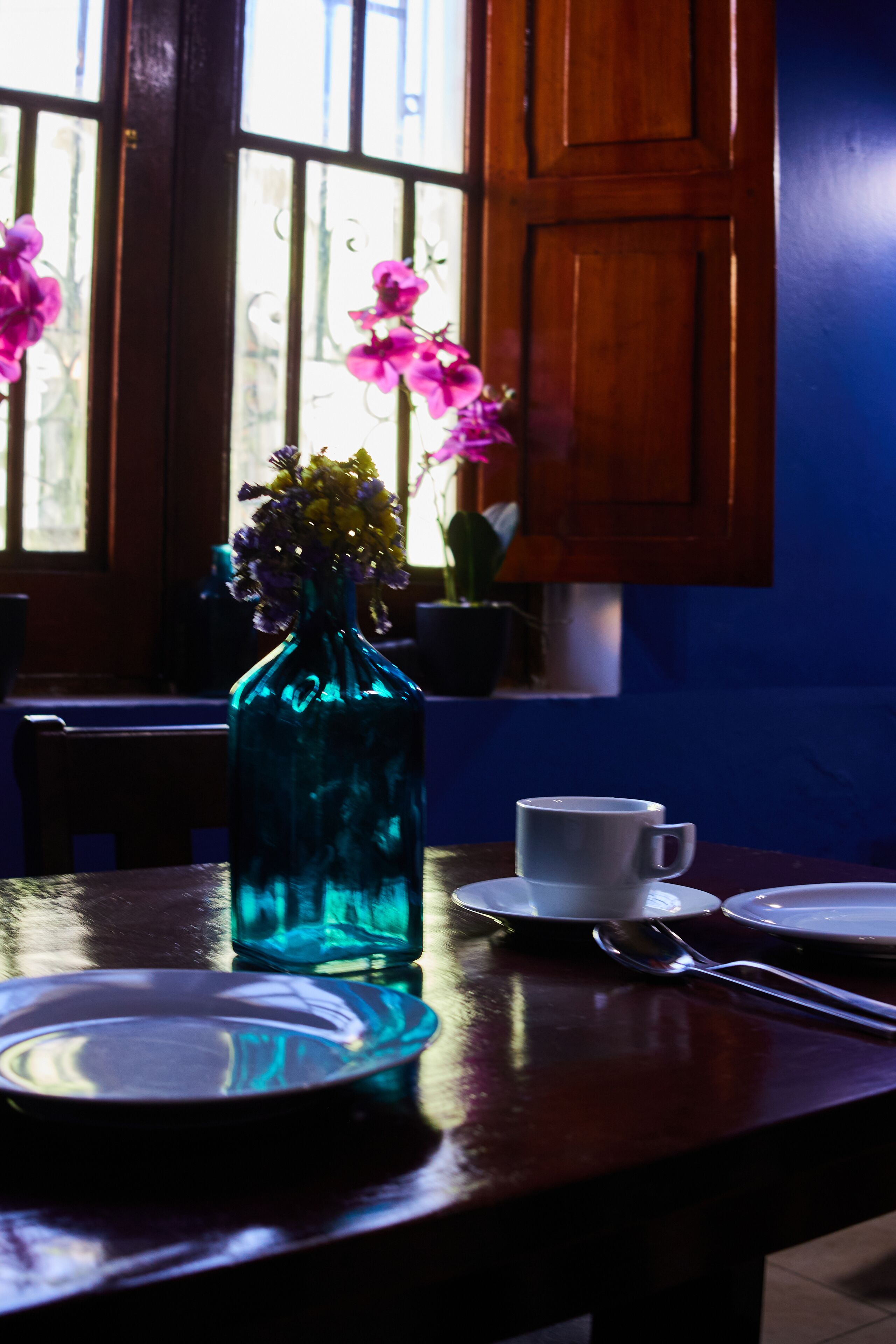 Una mesa con un jarrón azul de vidrio que contiene flores, una taza blanca y una cuchara.