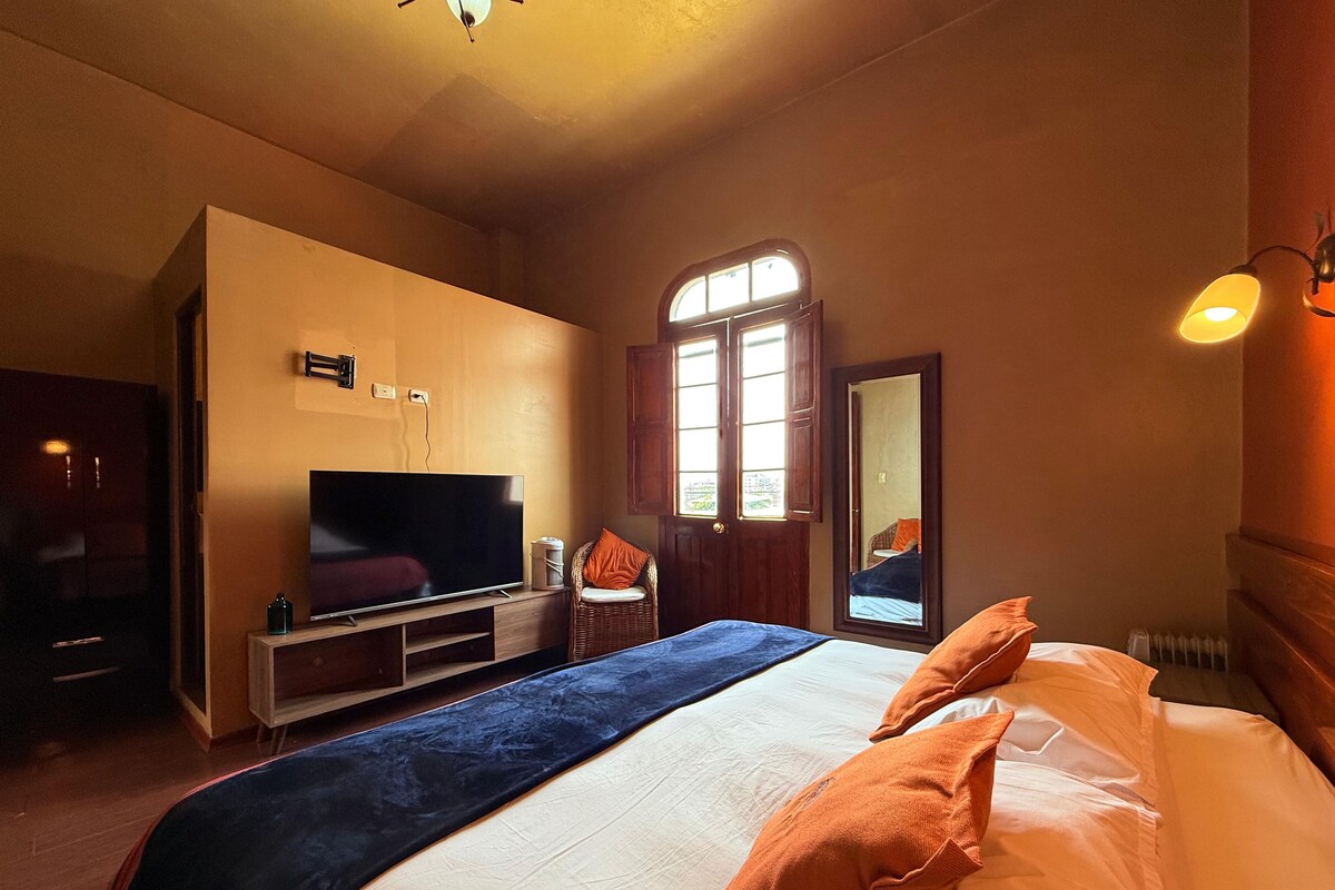 A hotel room with a large bed, a flat-screen TV, a wicker chair, and a window with curtains.