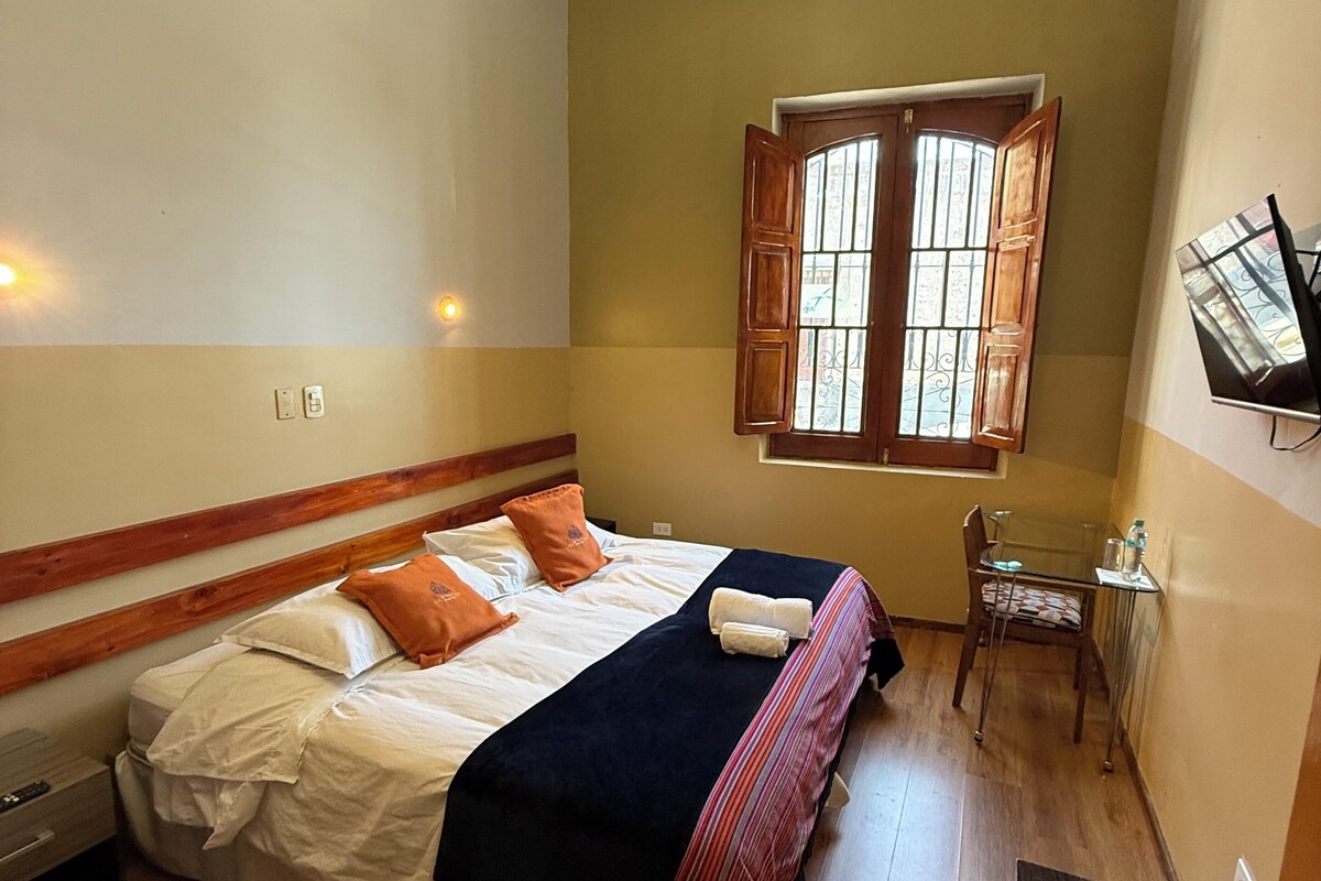 A bedroom with two beds, a window with wooden shutters, a small table, and a TV mounted on the wall.