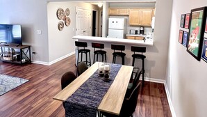 A modern kitchen with wooden cabinets, a white refrigerator, and a microwave. There is a dining area with a wooden table and black chairs. - 4 Bed 4 Bath Condo Near Campus! (Auburn)