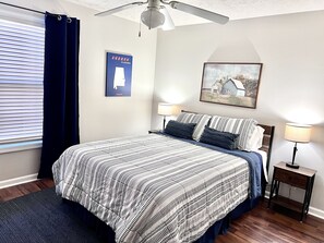 Une chambre à coucher comprenant un lit, deux tables de chevet, un ventilateur de plafond, une fenêtre avec des rideaux, un tableau accroché au mur et une carte encadrée.