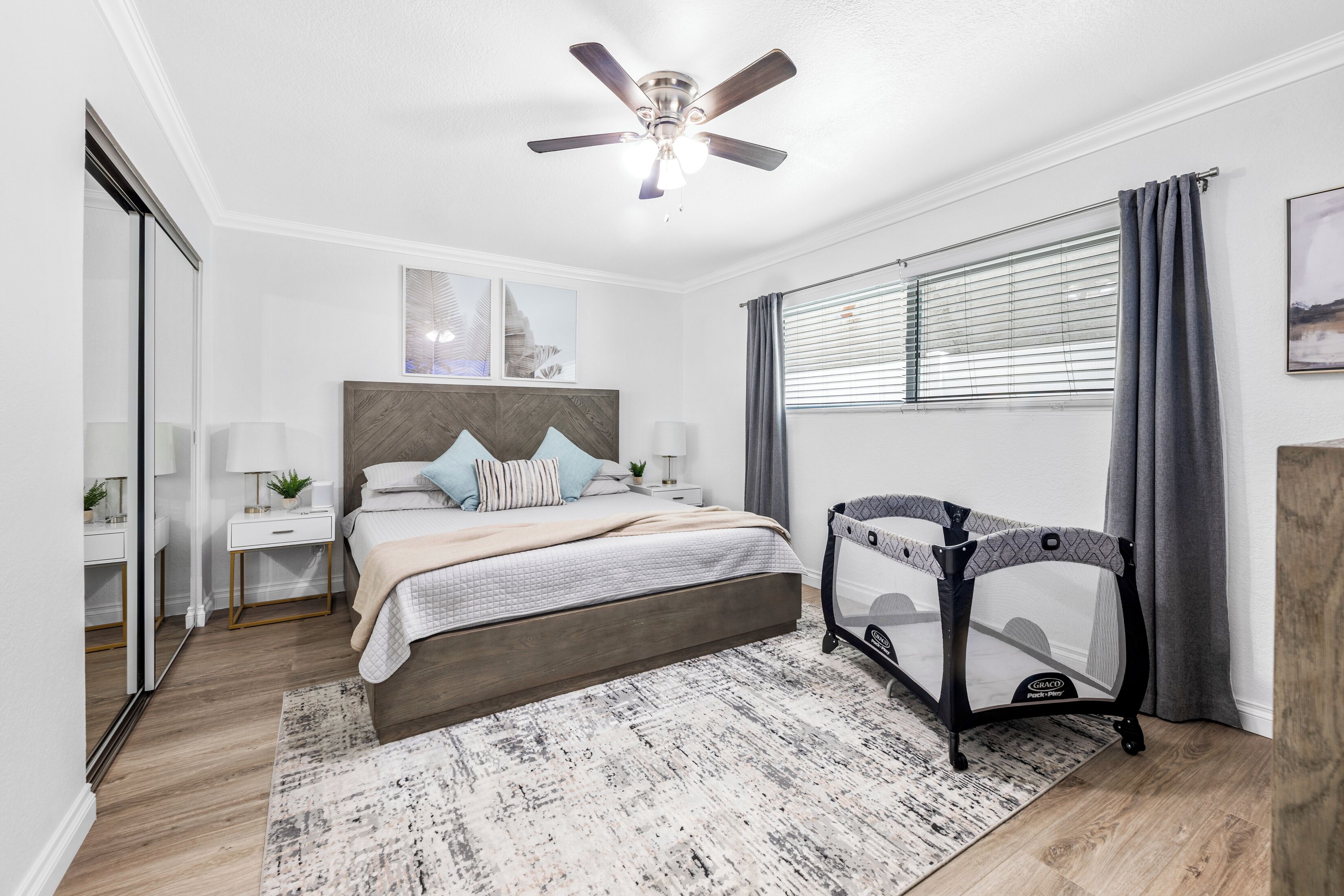 Ein Schlafzimmer mit einem Bett, Nachttischen, einem Deckenventilator, einem Babybett und einem Fenster mit Vorhängen.