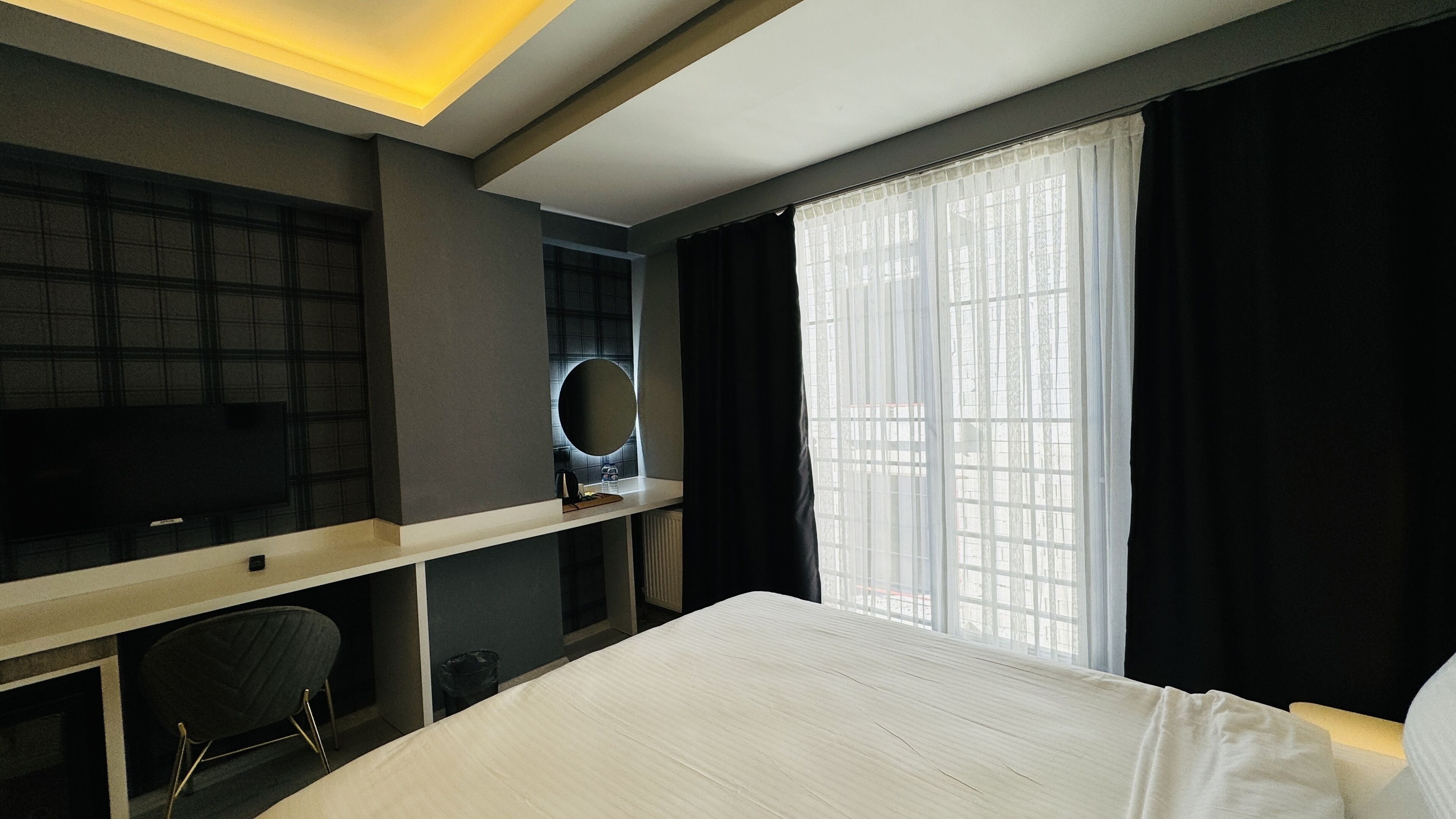 A hotel room with a bed, a desk with a chair, a mirror, and a window with sheer curtains.