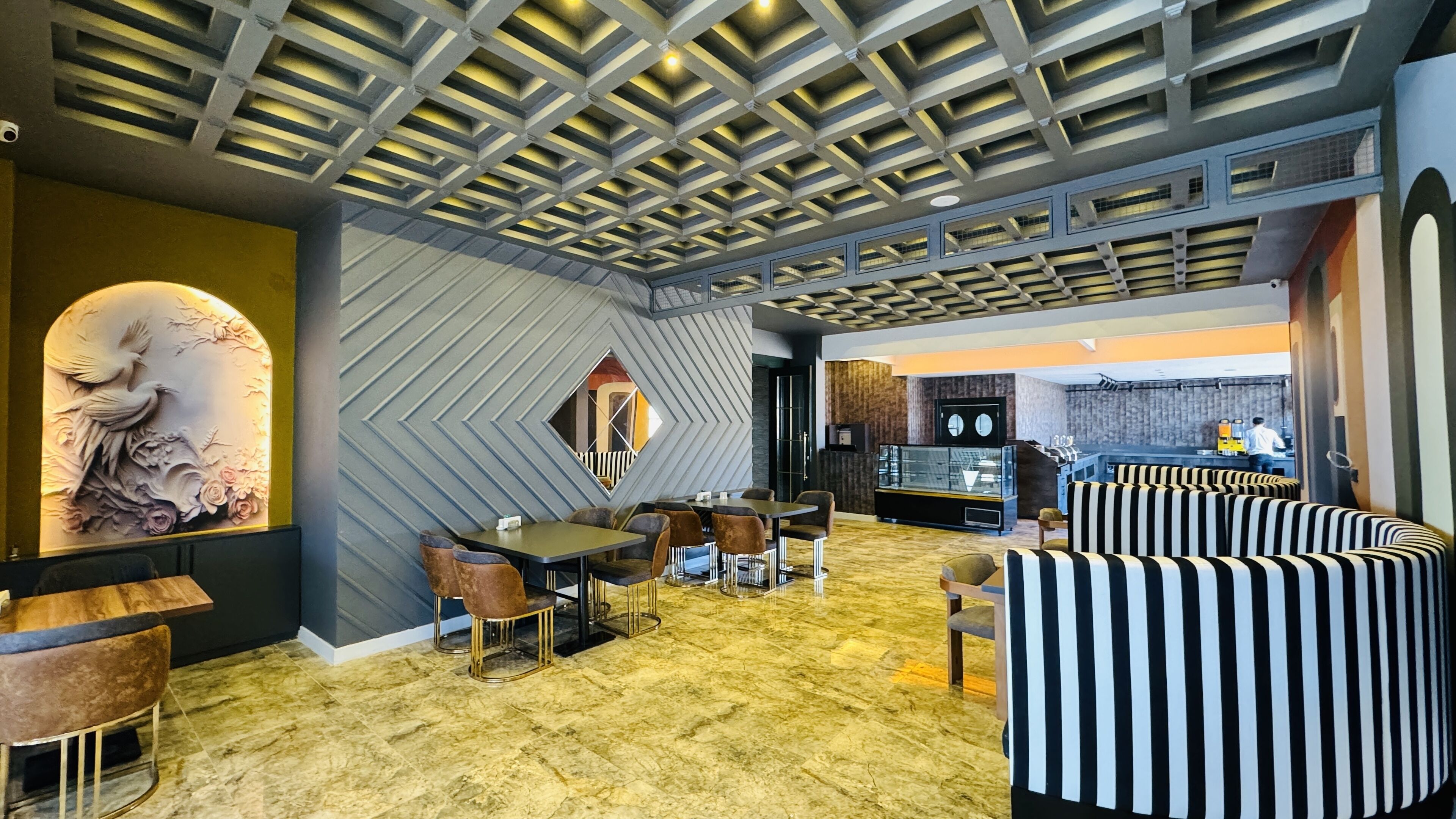 A modern interior with a geometric ceiling design, a dining area with tables and chairs, a bar area, and a seating area with striped sofas.