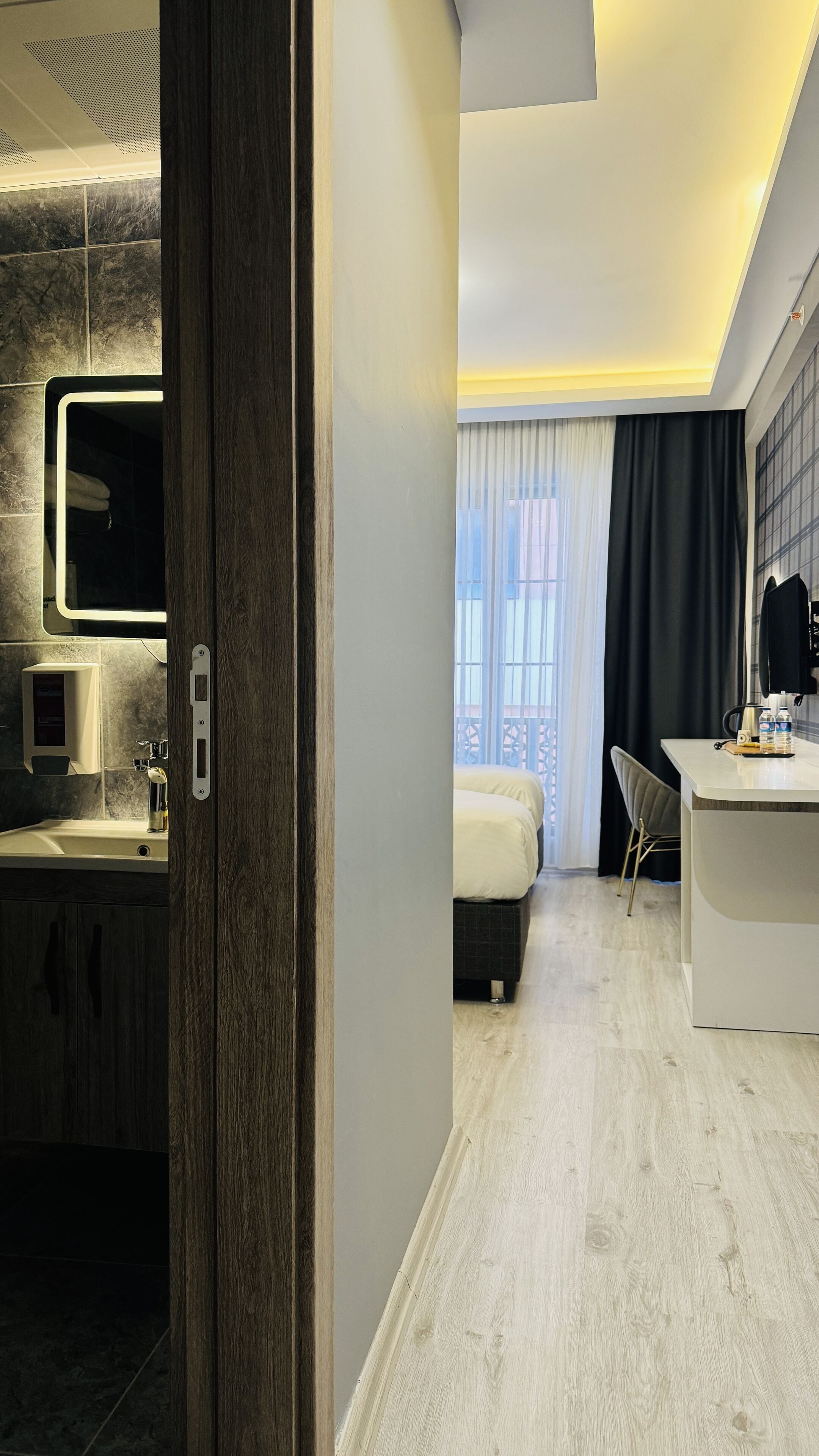 Una camera d'albergo moderna con una porta in legno, un bagno con uno specchio e un lavandino, un letto con una testiera nera, una scrivania con una sedia e una finestra con tende.