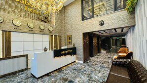 Zona de recepción de un hotel con mostrador de recepción, relojes que muestran la hora en Moscú, Estambul, Nueva York y Londres, una araña y un área de descanso con sofá y sillones.