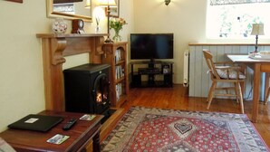 A cozy living room with a fireplace, a television, a dining area, and a large window with a view of greenery. - The Granary - Lanjeth (St Austell)