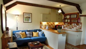 A cozy living room with a blue sofa, wooden coffee table, and a kitchen area with a tiled backsplash and wooden cabinets. - The Stables - Lanjeth (St Austell)