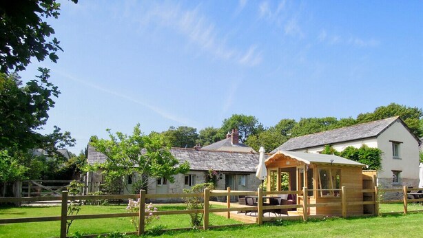 A rustic wooden structure with a roof, surrounded by a wooden fence, in a lush green field with a stone building in the background.