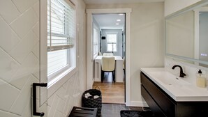 A modern bathroom with a white sink, black faucet, and a large mirror. There is a window with blinds on the left and a trash bin in the corner.