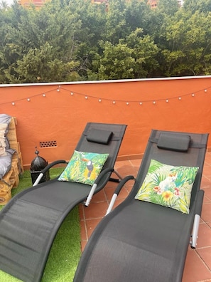 Two lounge chairs with floral cushions, a small table, and a string of lights against an orange wall.