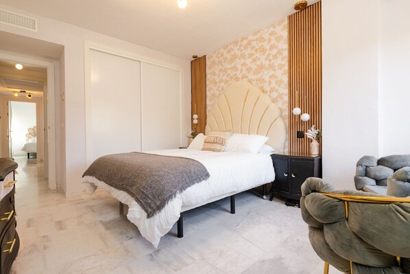 A bedroom with a bed, bedside table, nightstand, and a chair.