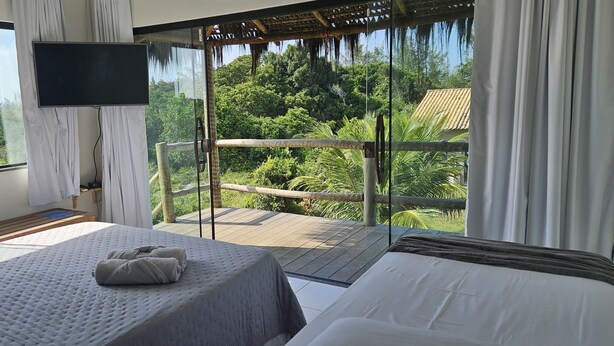 A bedroom with a large window, a bed, a TV, and a balcony with a view of the forest. - Kite Nomade (Arraial do Cabo)