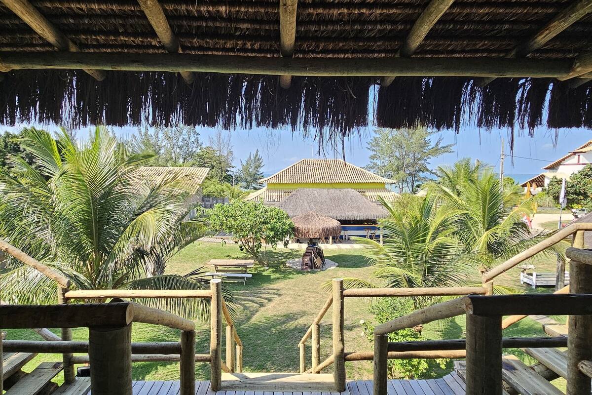 A wooden deck with a thatched roof, overlooking a pool area with palm trees and a building with a striped roof.