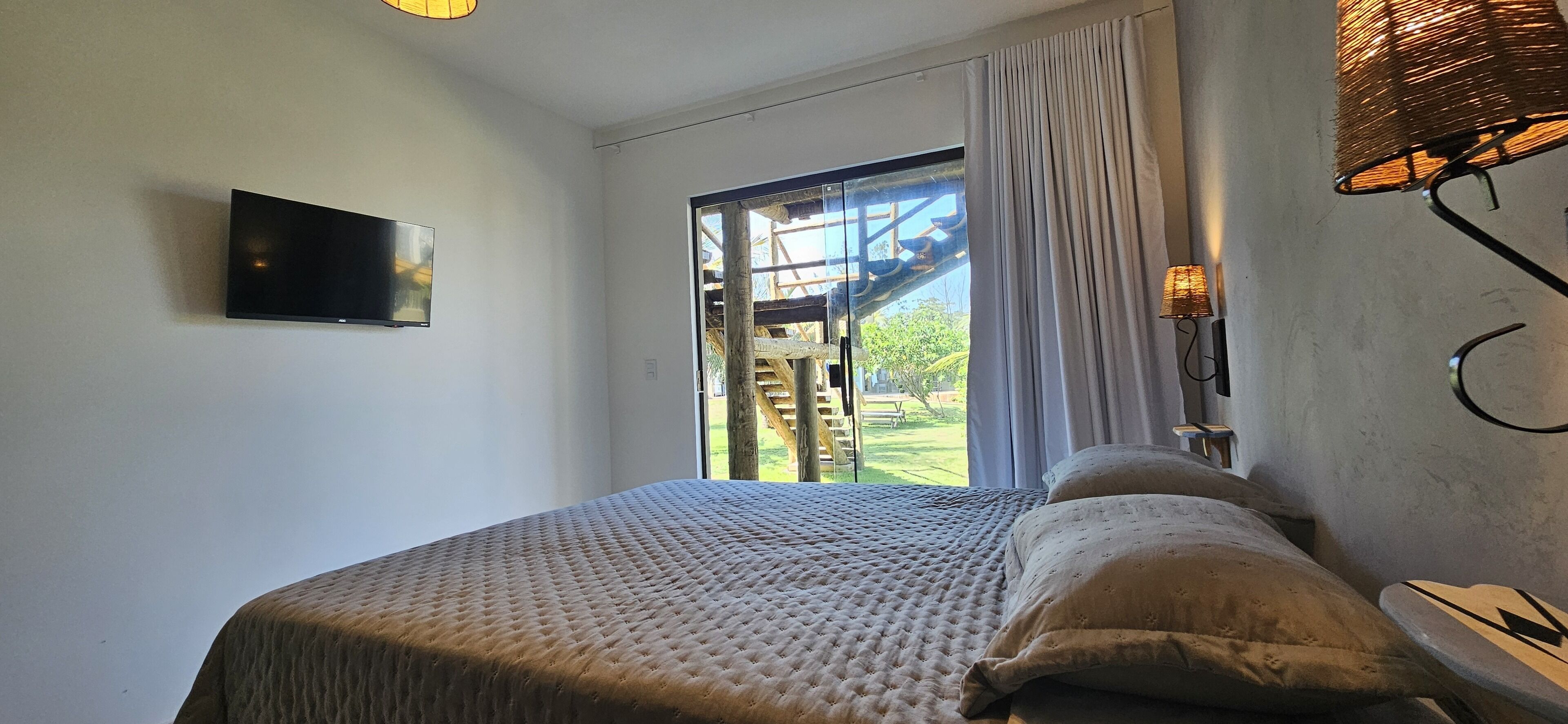 Un dormitorio con cama, televisor, ventana con cortinas y vista a un espacio exterior.