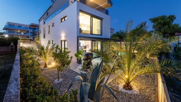 Une maison moderne à deux étages, dotée d’un toit plat, de grandes fenêtres et d’un espace extérieur bien aménagé, avec des plantes et un barbecue.