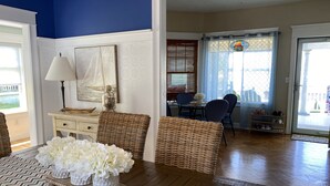 A living room with wicker furniture, a dining area with blue chairs, and a view of the outside through a window with a stained glass design. - BELLE of the BEACH: 30 ft to gorgeous PRIVATE BEACHFRONT home SLEEPS up to 13 (Wareham)
