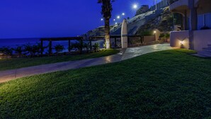 Un camino bien iluminado que lleva a una playa con una palmera y una tabla de surf, rodeado por un edificio moderno con balcones y luces.