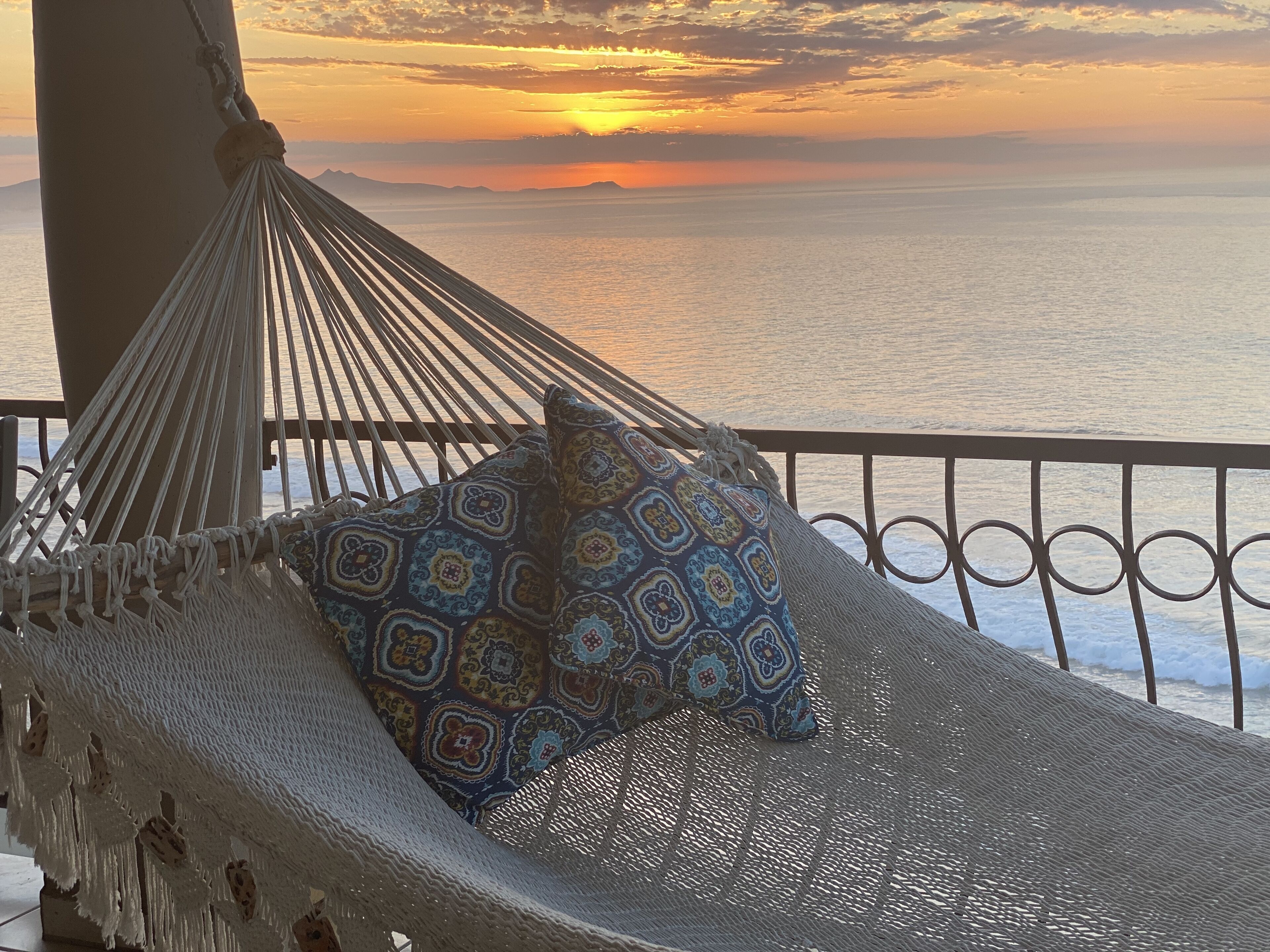 Een hangmat met een gekleurd kussen op een balkon met uitzicht op zee bij zonsondergang.