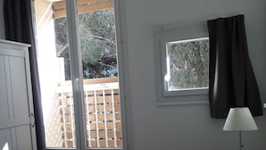 Quarto com cama, janela com vista para um deque de madeira e árvores, luminária e guarda-roupa.