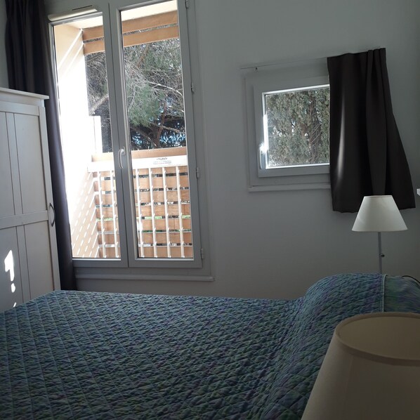 Un dormitorio con cama, ventana con vista a una terraza de madera y árboles, lámpara y armario.
