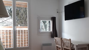 Um quarto com piso de madeira, um sofá verde, uma mesa de jantar com cadeiras, uma TV fixada na parede e uma janela com vista para árvores.
