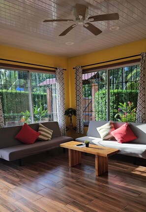 A room with a ceiling fan, two sofas with cushions, a wooden coffee table, and large windows with curtains.