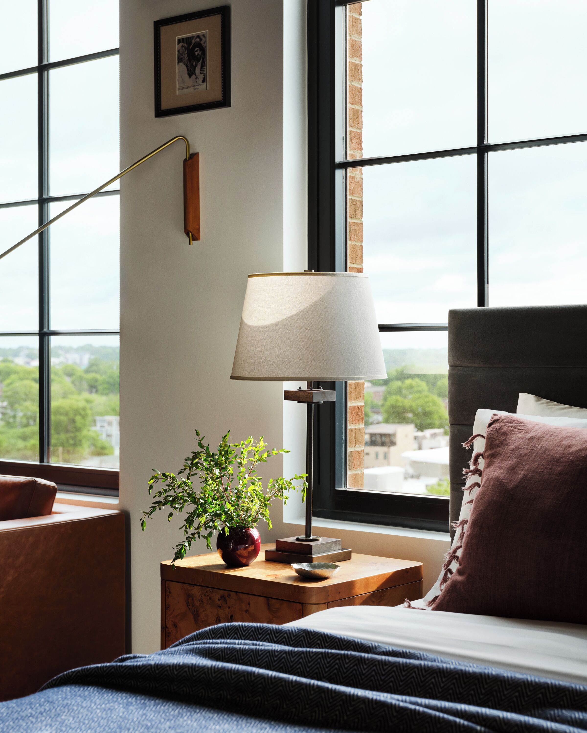 A bedroom with a bed, a bedside table with a lamp, a plant, and a window with a view of greenery.