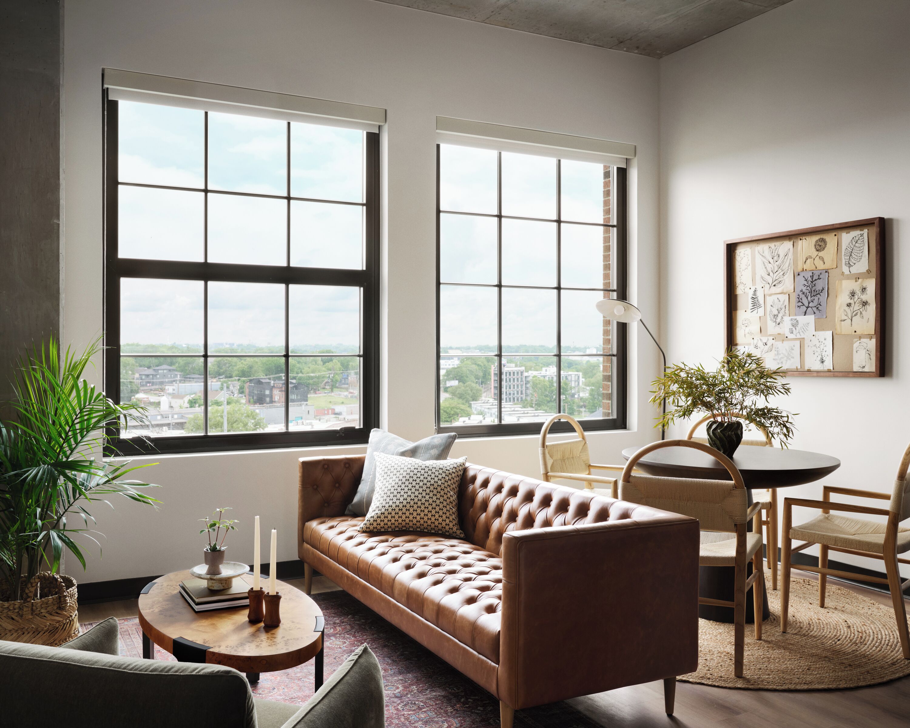 A modern living room with a leather sofa, a round coffee table, and a dining table with chairs. There are large windows with a city view, a potted plant, and a wall-mounted art piece.
