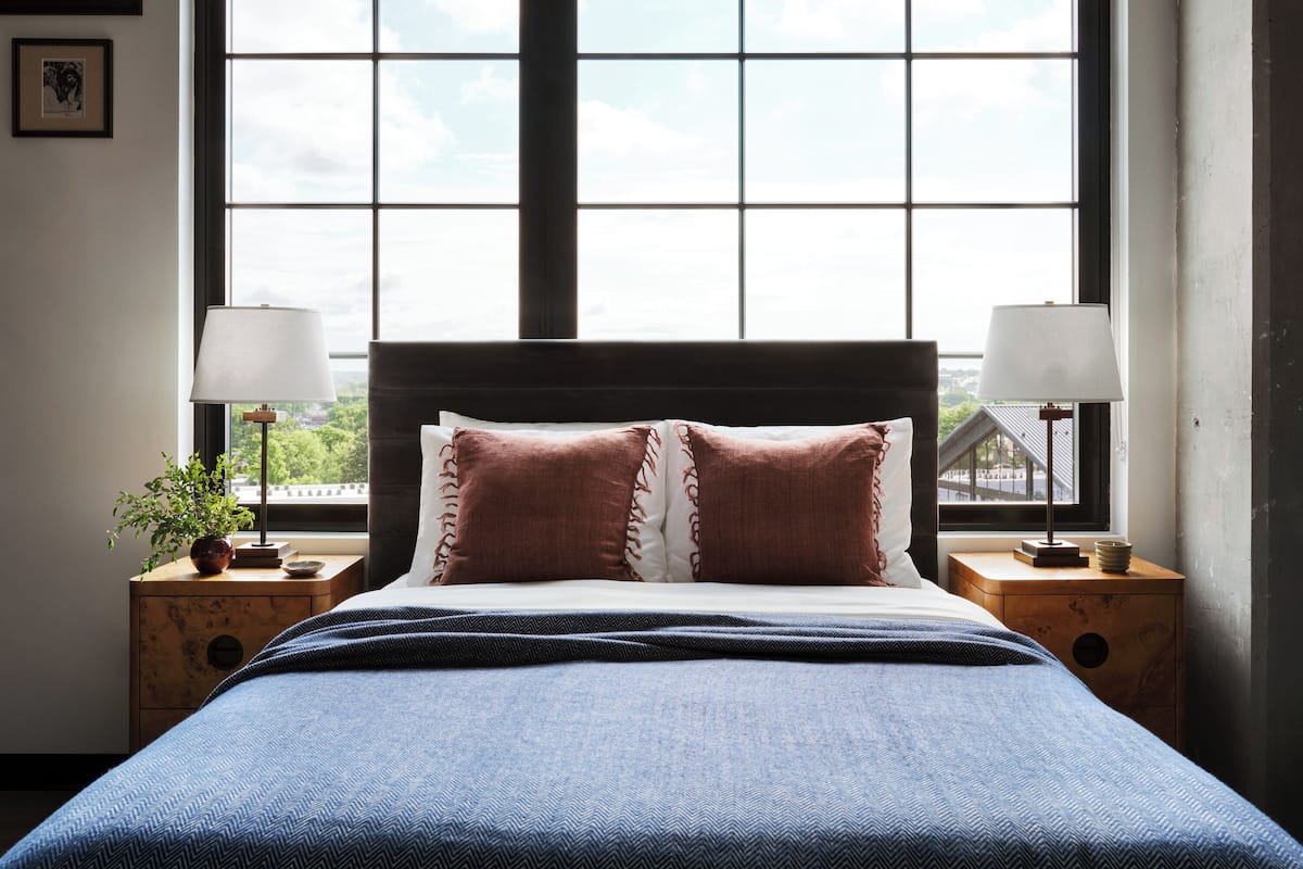 Een slaapkamer met een groot raam, een bed met een blauwe sprei, twee nachtkastjes met lampen, en uitzicht op groen buiten.