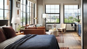 A modern bedroom with a large bed, a sofa, a desk, and a view of the city through large windows.