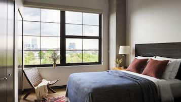 Un dormitorio con un ventanal grande, una cama con una colcha azul, una mesita de noche con una lámpara y una silla junto a la ventana.