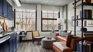A modern living room with a sofa, ottoman, and a bookshelf filled with books. There is a window with a view of trees and a lamp on a side table.