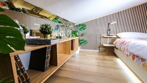 A modern bedroom with a wooden floor, a bed with a colorful blanket, a desk with books and a lamp, and a wall with vertical wooden slats and green plants. - Flytosuite aeroporto attico mansarda  (Calderara di Reno)