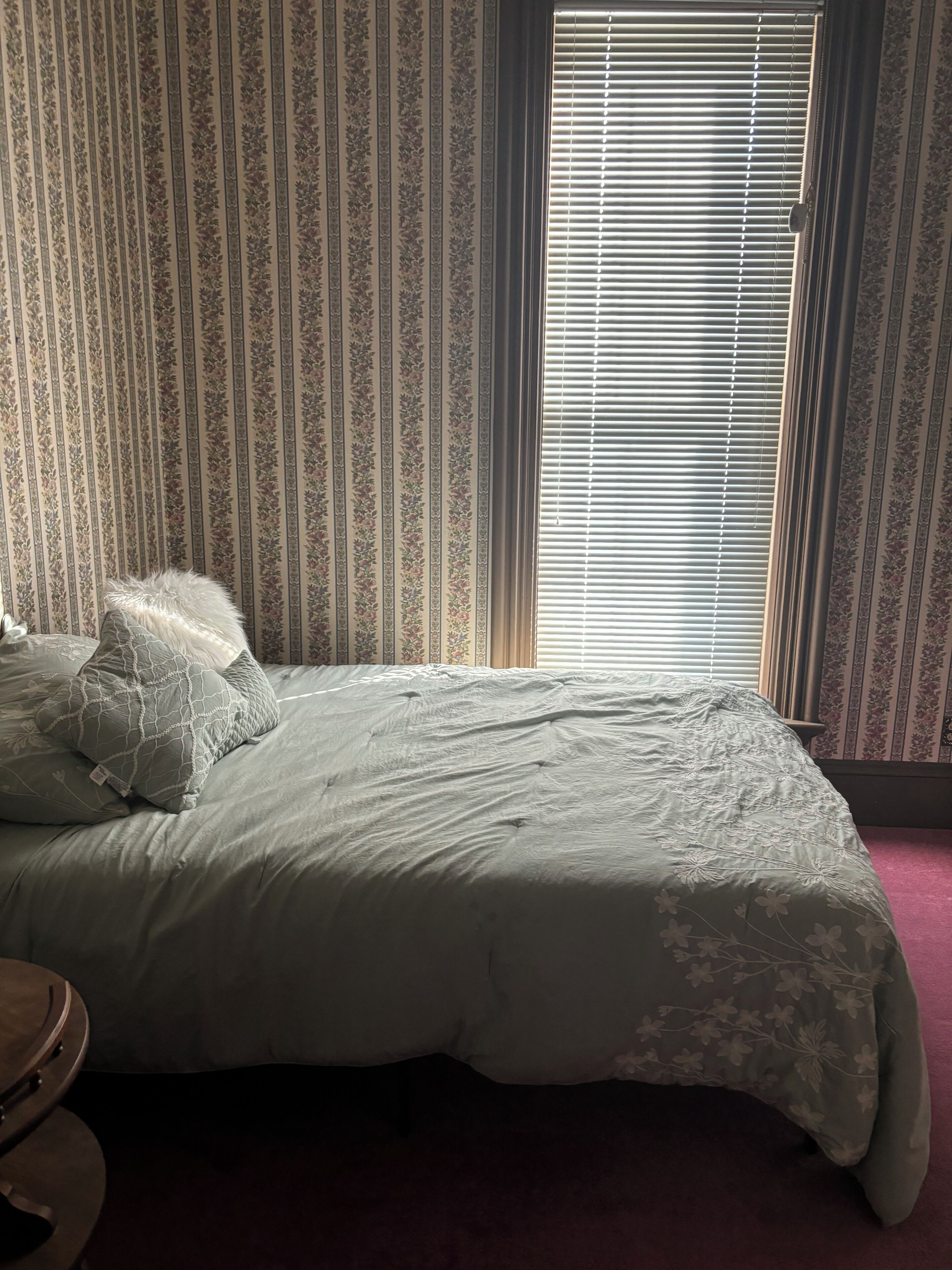 A bedroom with a bed, pillows, a window with blinds, and striped wallpaper.