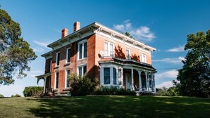 Une grande maison à deux étages, construite en briques, dotée d’un porche qui fait le tour de la maison et d’une pelouse bien entretenue.