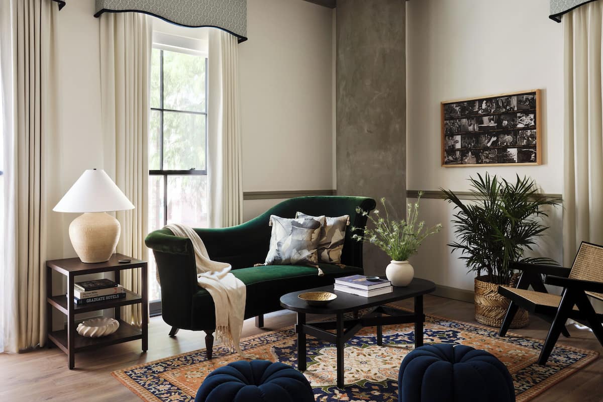 Een woonkamer met een groene bank, een salontafel, een lamp, een boekenkast, een groot raam met gordijnen, een kamerbreed tapijt met patroon en een ingelijste foto aan de muur.