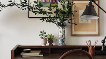 A wooden desk with a woven chair, a vase with plants, books, and framed artwork on the wall.