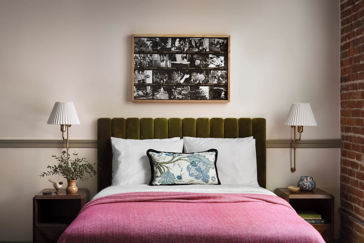 Een slaapkamer met een groen gestoffeerd hoofdeinde, een roze sprei en twee nachtkastjes met lampen. Daarboven hangt een ingelijste collage.