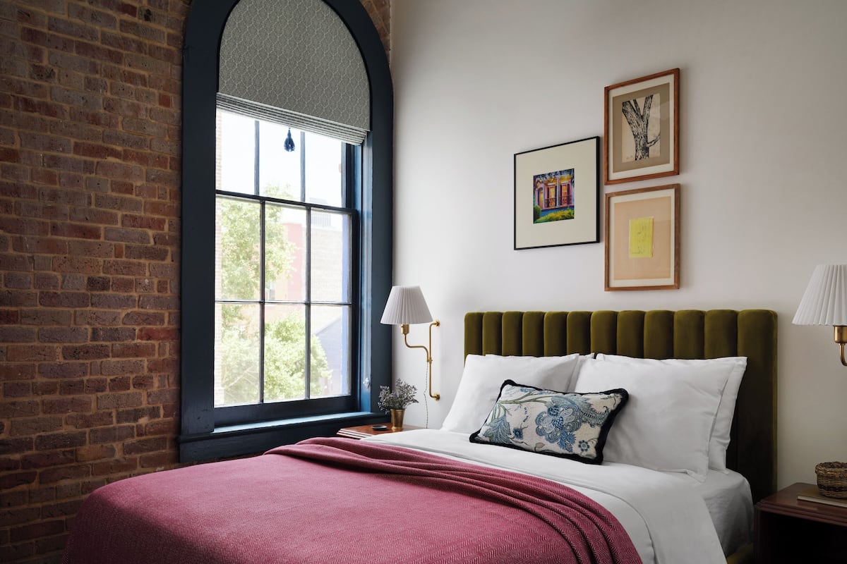 Een slaapkamer met een bakstenen muur, een groot raam, een bed met een groene hoofdeinde en twee wandlampen.