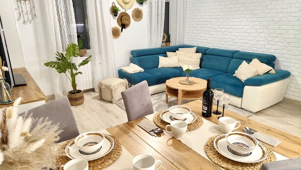 A modern living room with a blue sofa, a dining table set with plates, cups, and cutlery. There are decorative hats on the wall and a dreamcatcher.