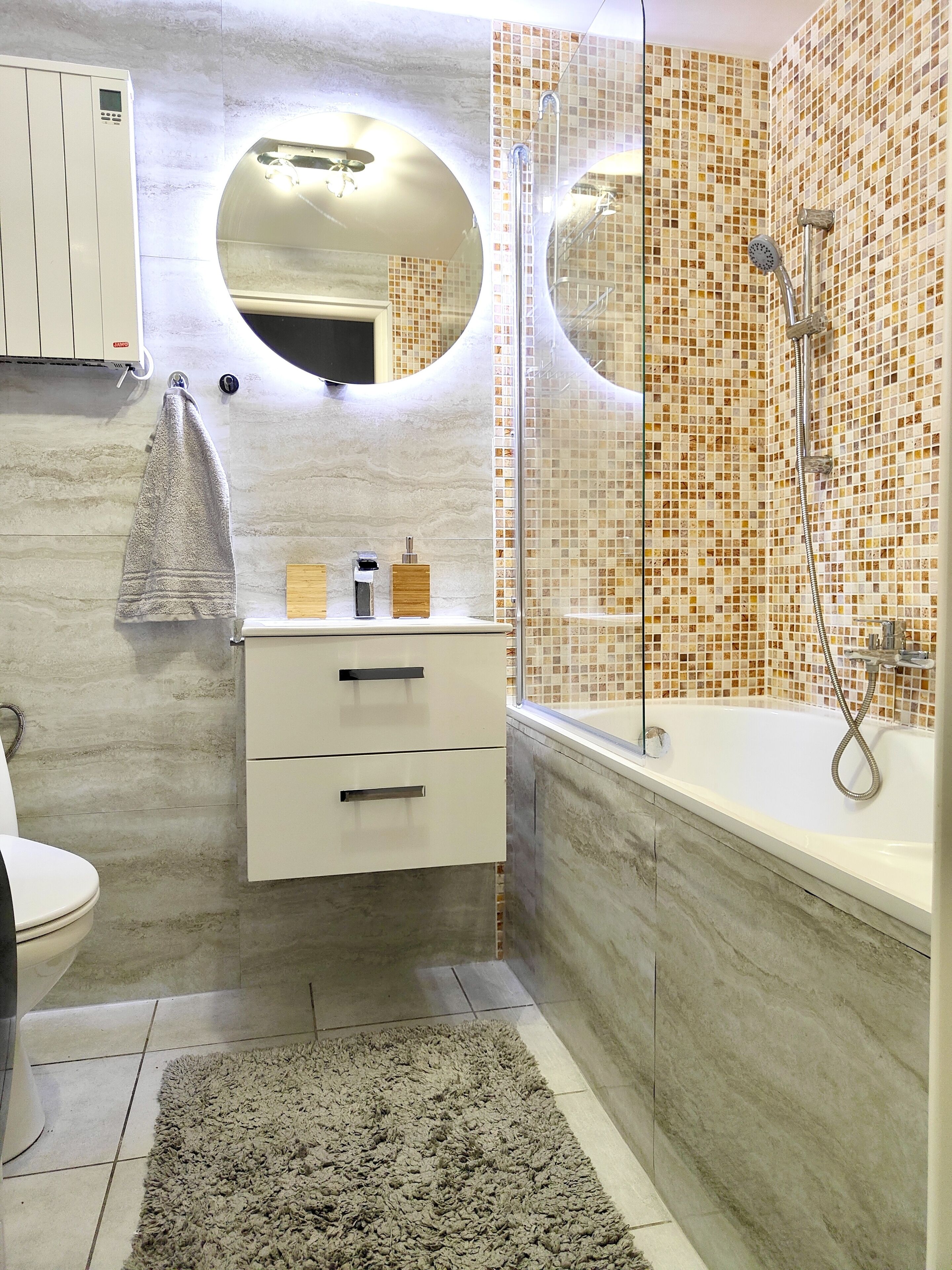 A modern bathroom with a glass-enclosed shower, a round mirror, a white vanity with two drawers, and a towel hanging on the wall.