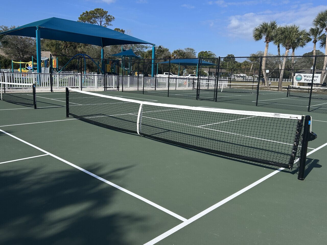 Una cancha de tenis con una red, un toldo azul y un letrero que dice 'Welcome Players!'.